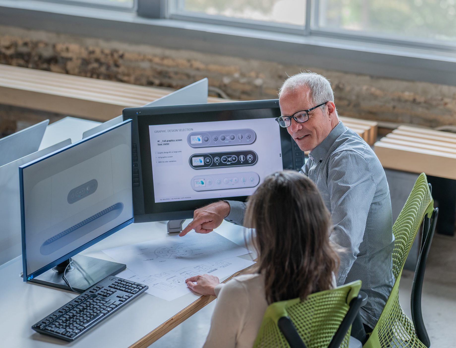 Two people sit at a desk with computer monitors, discussing a technical diagram shown on the screens and paper. As they talk in a modern office with large windows, they review a stratégie de design au Québec for an upcoming project.