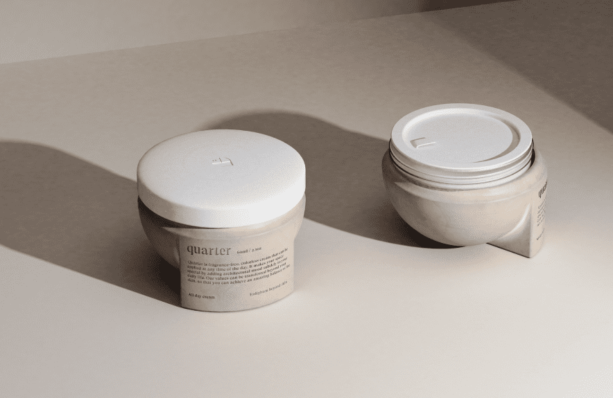 Two beige jars of Quartier moisturizer, designed with expertise from an agence de design industriel à Montréal, sit on a neutral surface—one upright and one on its side—casting soft shadows in minimal lighting.