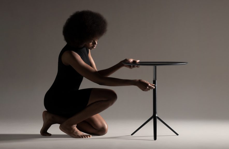 A person with an afro hairstyle, dressed in a black outfit, kneels barefoot on the floor while assembling a sleek round table—embodying une stratégie de design au Québec—against a neutral background.