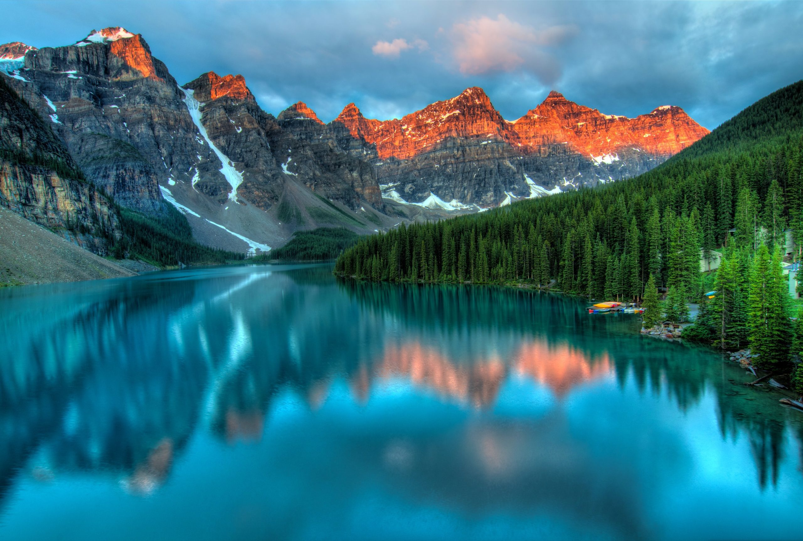 A serene turquoise lake reflects pine trees and rugged mountain peaks bathed in orange sunlight, under a cloudy sky—an ideal scene for inspiration at une agence d'ingénierie mécanique à Montréal or for developing une stratégie de design au Québec.