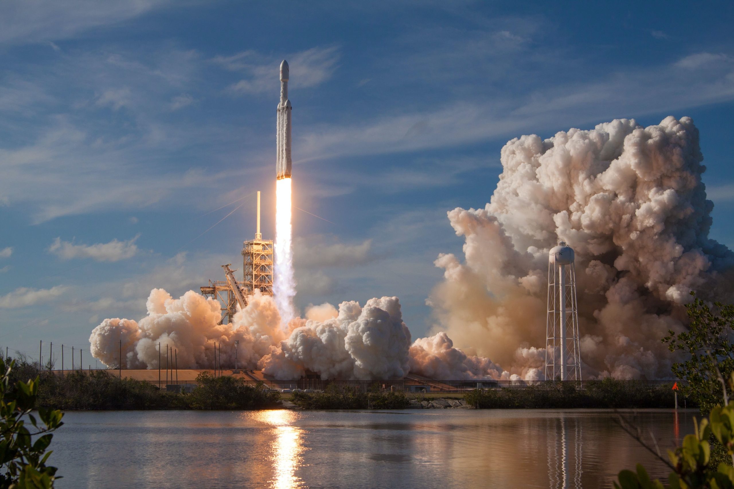 A rocket launches into the blue sky, leaving a fiery trail and billowing exhaust, as a body of water reflects the scene—showcasing innovation worthy of an agence de développement de produits à Montréal.