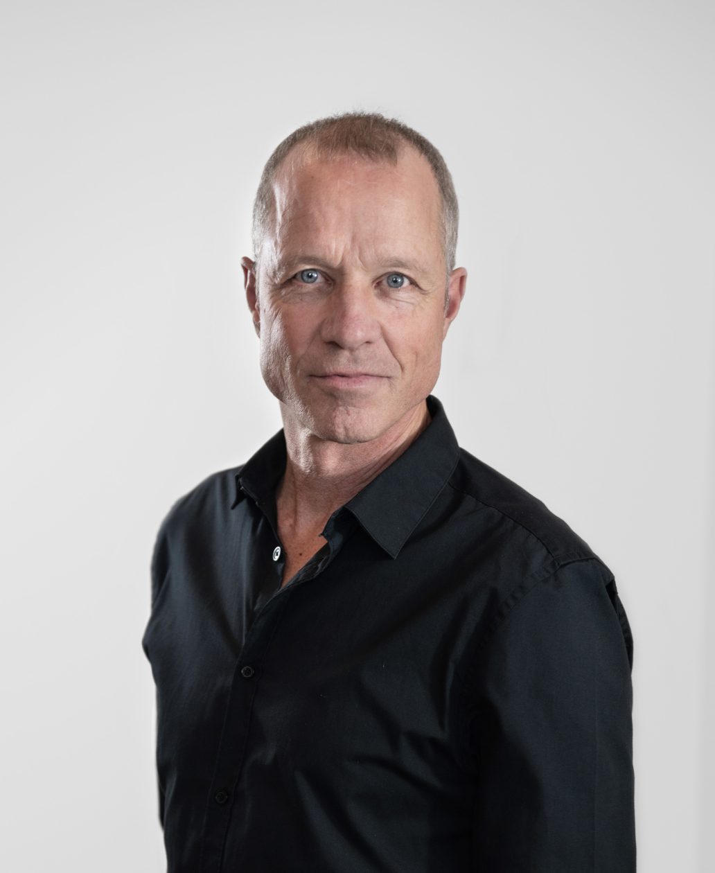 A middle-aged man with short gray hair and blue eyes, wearing a black button-up shirt, stands in front of a plain white background, embodying the confidence of an expert from an agence de design industriel à Montréal.