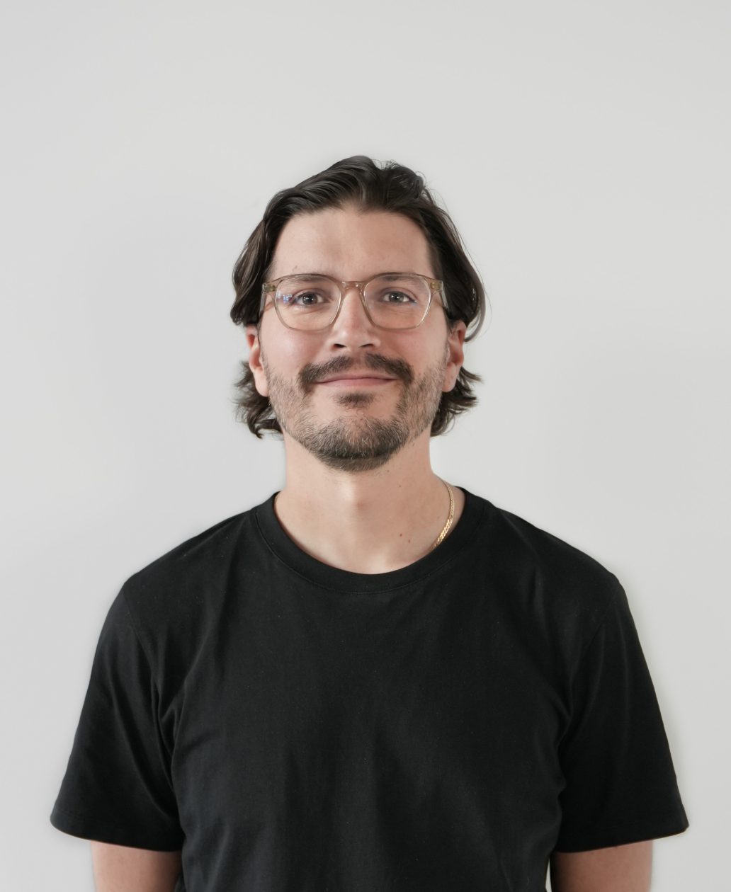 A man with medium-length dark hair, glasses, and a short beard is wearing a black t-shirt and standing in front of a plain white background, smiling slightly at the camera—an ideal image for an agence de développement de produits à Montréal.