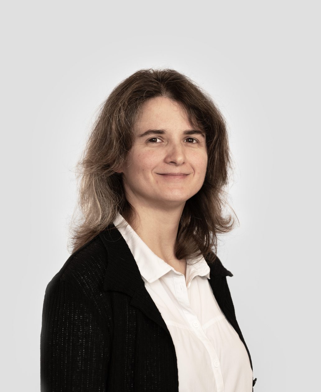A woman with shoulder-length brown hair, wearing a white shirt and a black jacket, smiles softly while facing slightly to the side—an approachable look perfect for an Agence de développement de produits à Montréal.
