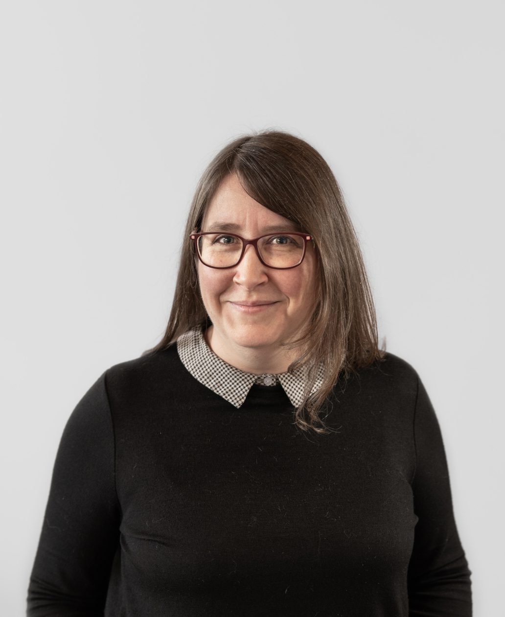 A person with straight brown hair and glasses, wearing a black sweater with a patterned collar, smiles slightly while standing against a plain light gray background, evoking the professionalism of an agence de développement de produits à Montréal.