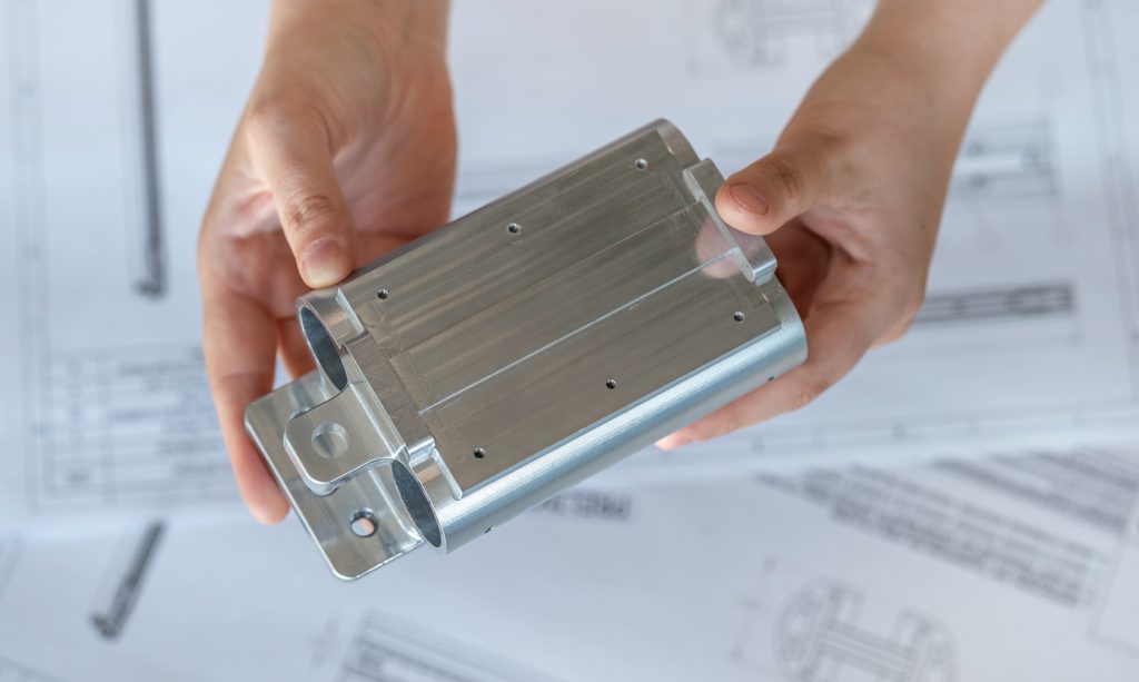 Une personne tient une pièce d'aluminium usinée avec des trous percés au-dessus de dessins techniques, mettant en évidence l'expertise d'une agence d'ingénierie mécanique à Montréal dans la livraison de composants mécaniques de précision.