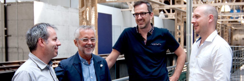 Quatre hommes se tiennent à l'intérieur d'une agence de développement de produits à Montréal, souriants et engagés dans une conversation. Vêtus de chemises et de vestes décontractées, ils se mêlent à une toile de fond de style industriel, faite de poutres et de tuyaux apparents.