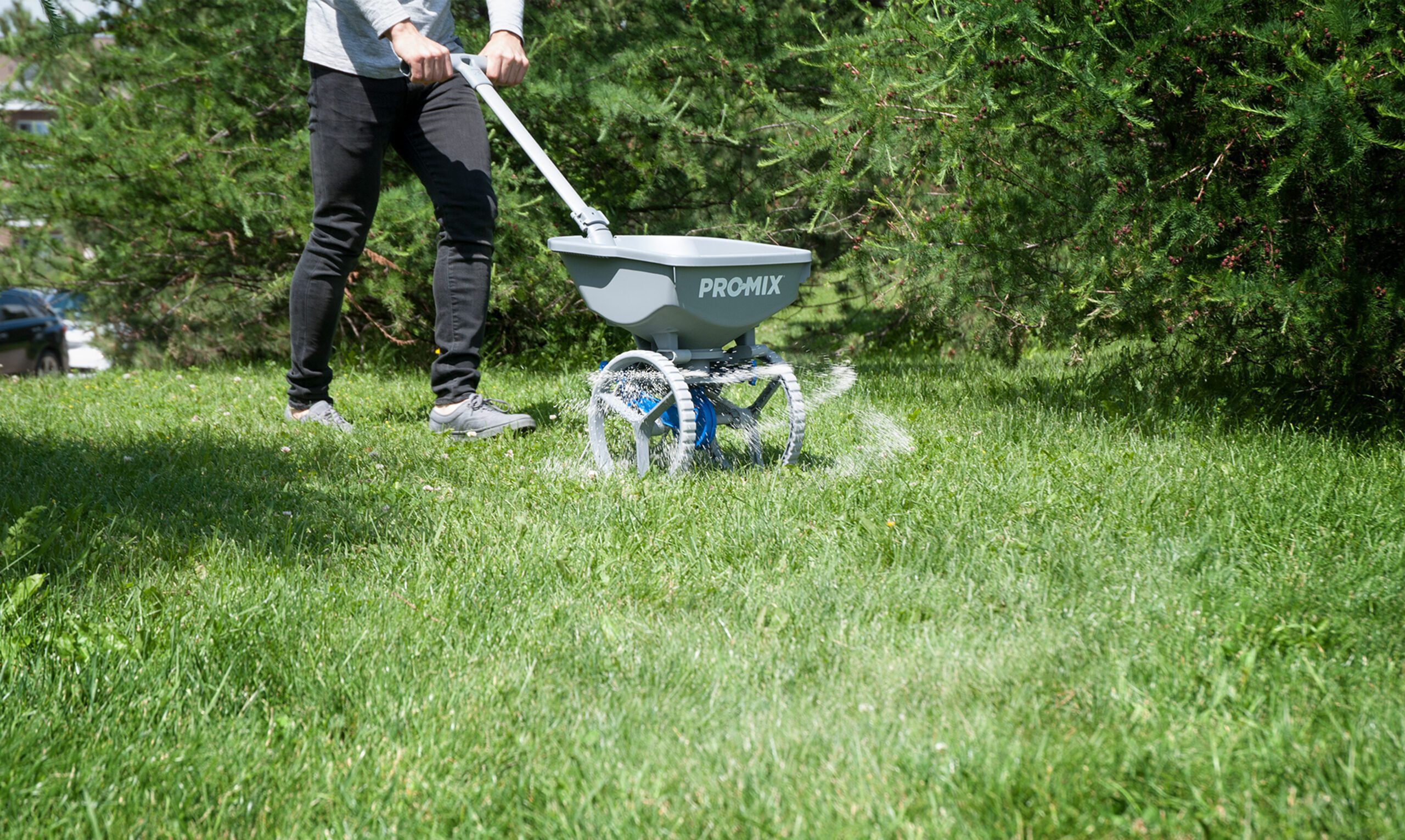 A person uses a PROMIX fertilizer spreader to distribute fertilizer on a green lawn, surrounded by bushes and trees—showcasing expertise inspired by une stratégie de design au Québec.