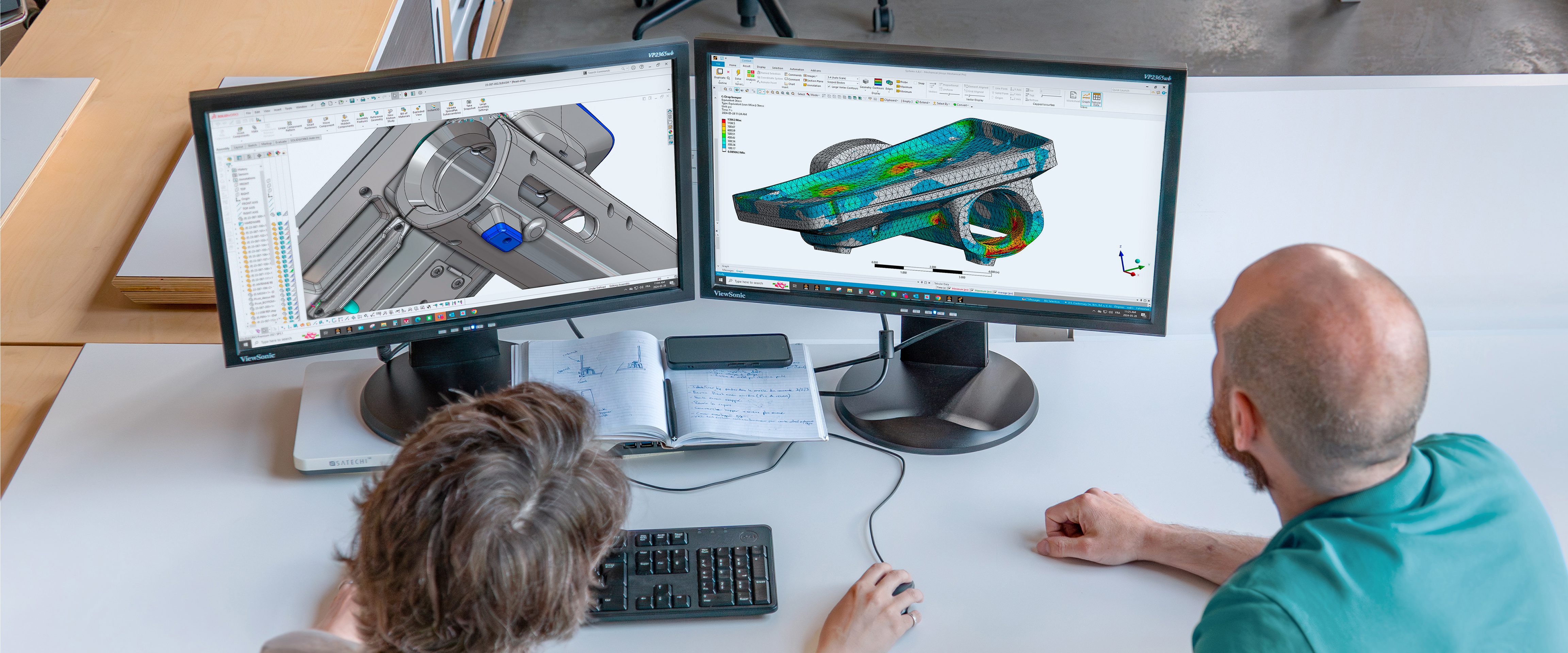 Deux personnes à un bureau, dans une agence d'ingénierie mécanique à Montréal, examinent des conceptions et des simulations mécaniques en 3D sur deux moniteurs, entourées d'un clavier, d'un ordinateur portable et d'un stylo.