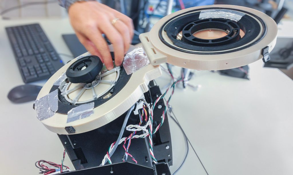 Une personne assemble ou répare un appareil avec des fils apparents et des composants circulaires en plastique à un bureau, reflétant l'expertise pratique que l'on retrouve souvent dans une agence de design industriel à Montréal. Un clavier et une souris d'ordinateur sont visibles à l'arrière-plan.