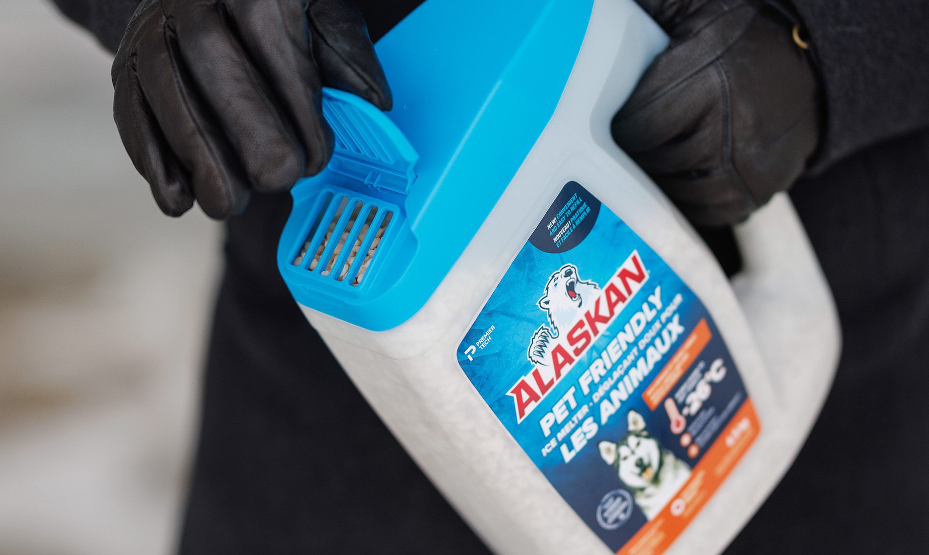 A person wearing black gloves holds a blue and white container of Alaskan Pet Friendly ice melter, designed for safe use around pets and effective in temperatures as low as -35°C, ideal for clients of an agence de design industriel à Montréal.