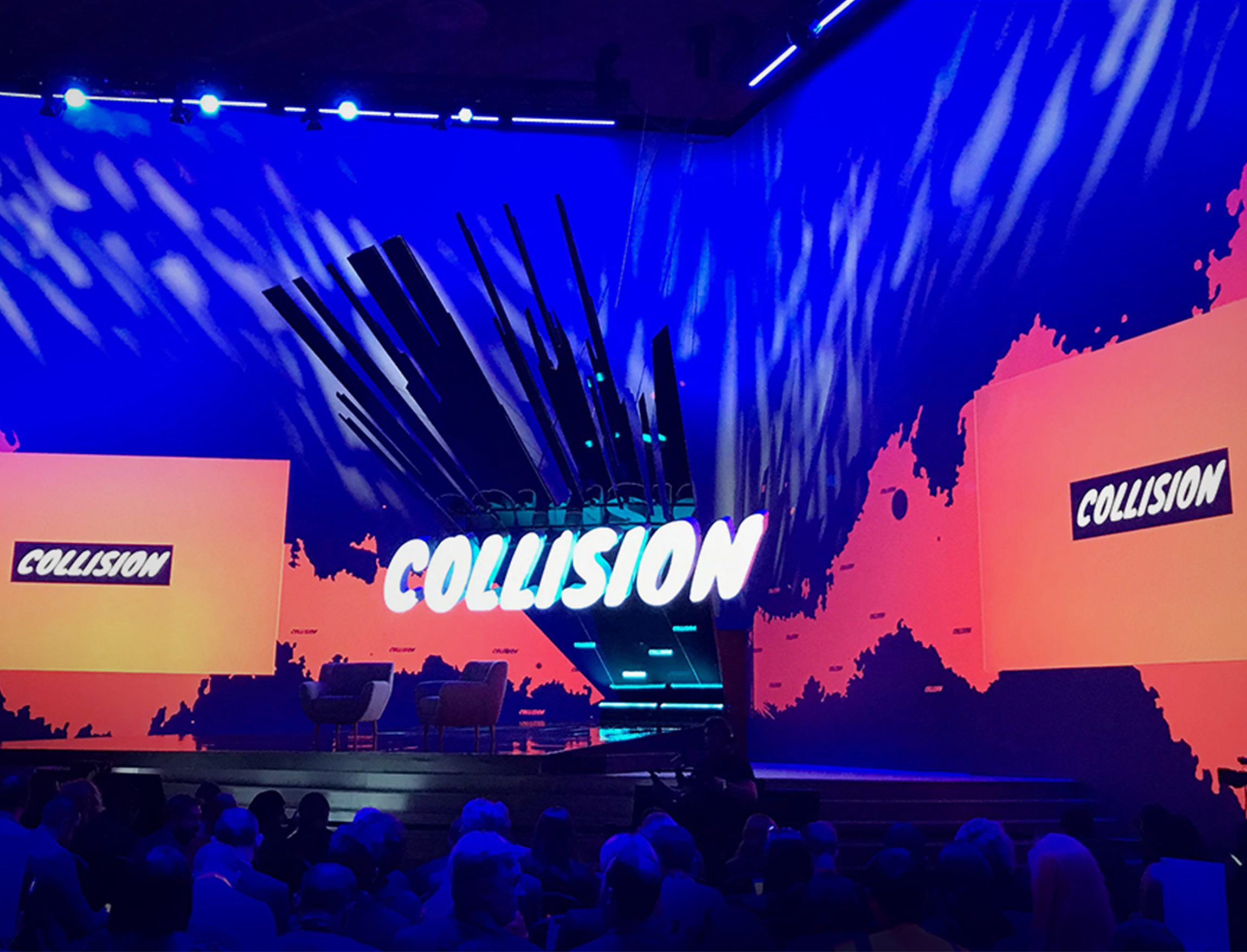 A brightly lit stage with “COLLISION” in large letters at the center, surrounded by abstract blue and orange lighting—perfect for an Agence de développement de produits à Montréal to showcase innovation. Two chairs and a table face the audience.