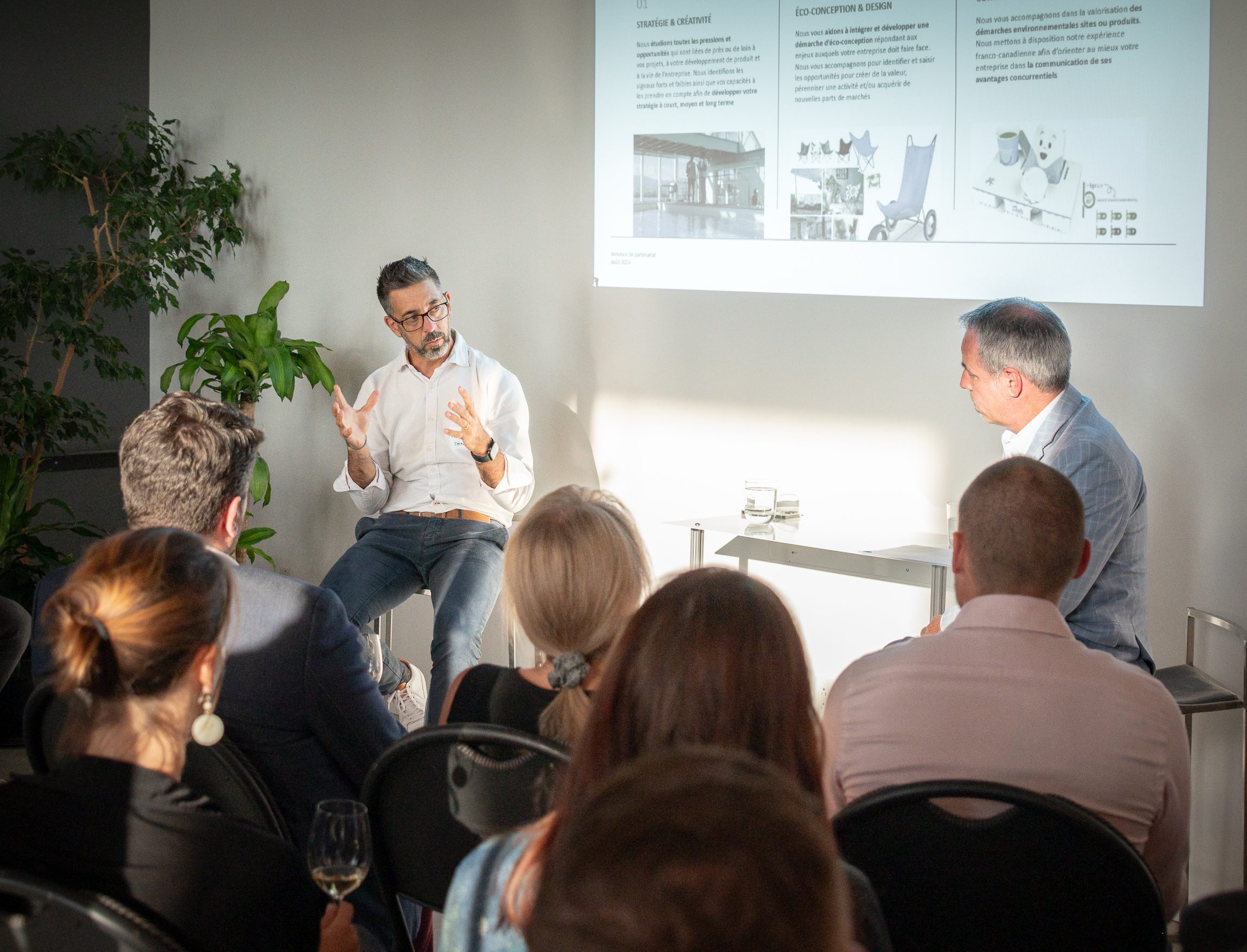 Un homme fait des gestes tout en parlant à un autre homme assis lors d'une présentation organisée par une agence de design industriel à Montréal, avec des diapositives projetées sur le mur. Le public écoute attentivement dans une salle bien éclairée et agrémentée de plantes.