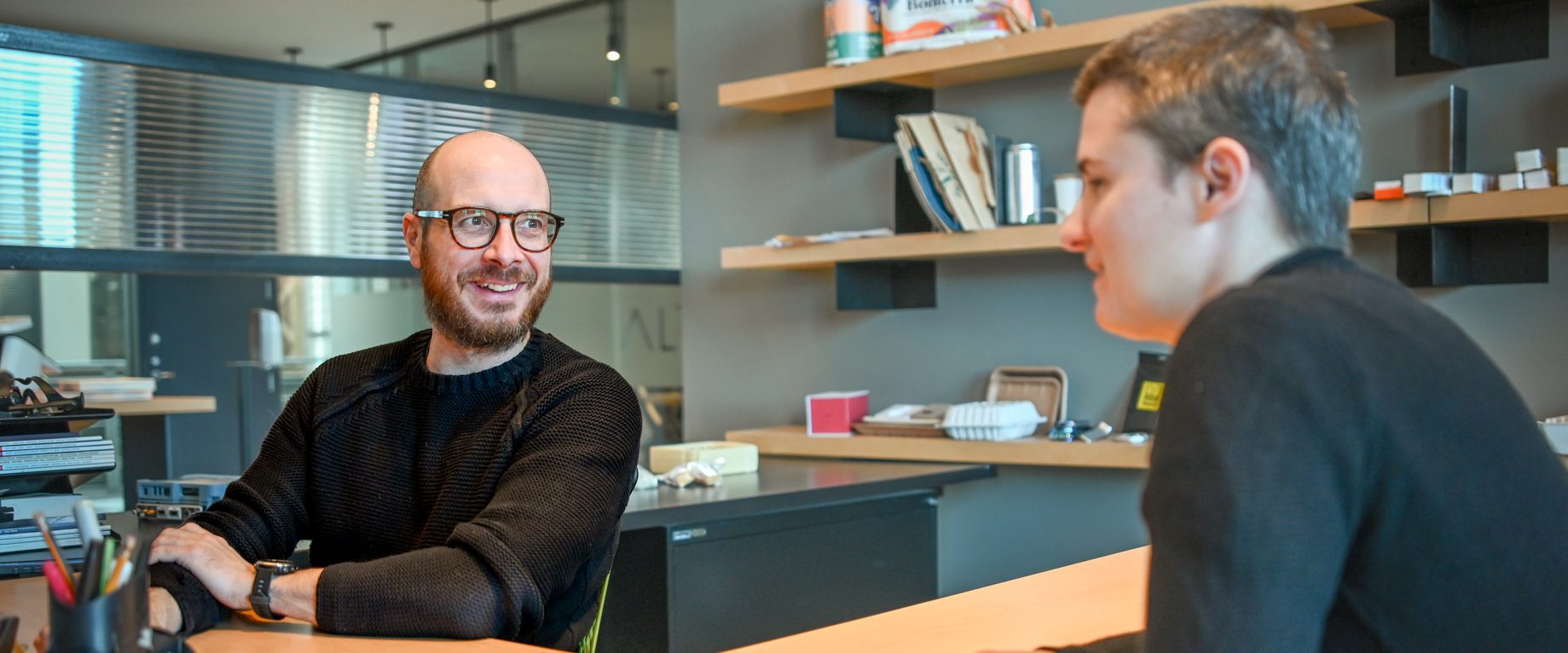 Deux personnes sont assises l'une en face de l'autre dans un bureau moderne, engagées dans une conversation. Une personne sourit et porte des lunettes ; des étagères et des fournitures de bureau sont visibles à l'arrière-plan de cette agence de design industriel à Montréal.