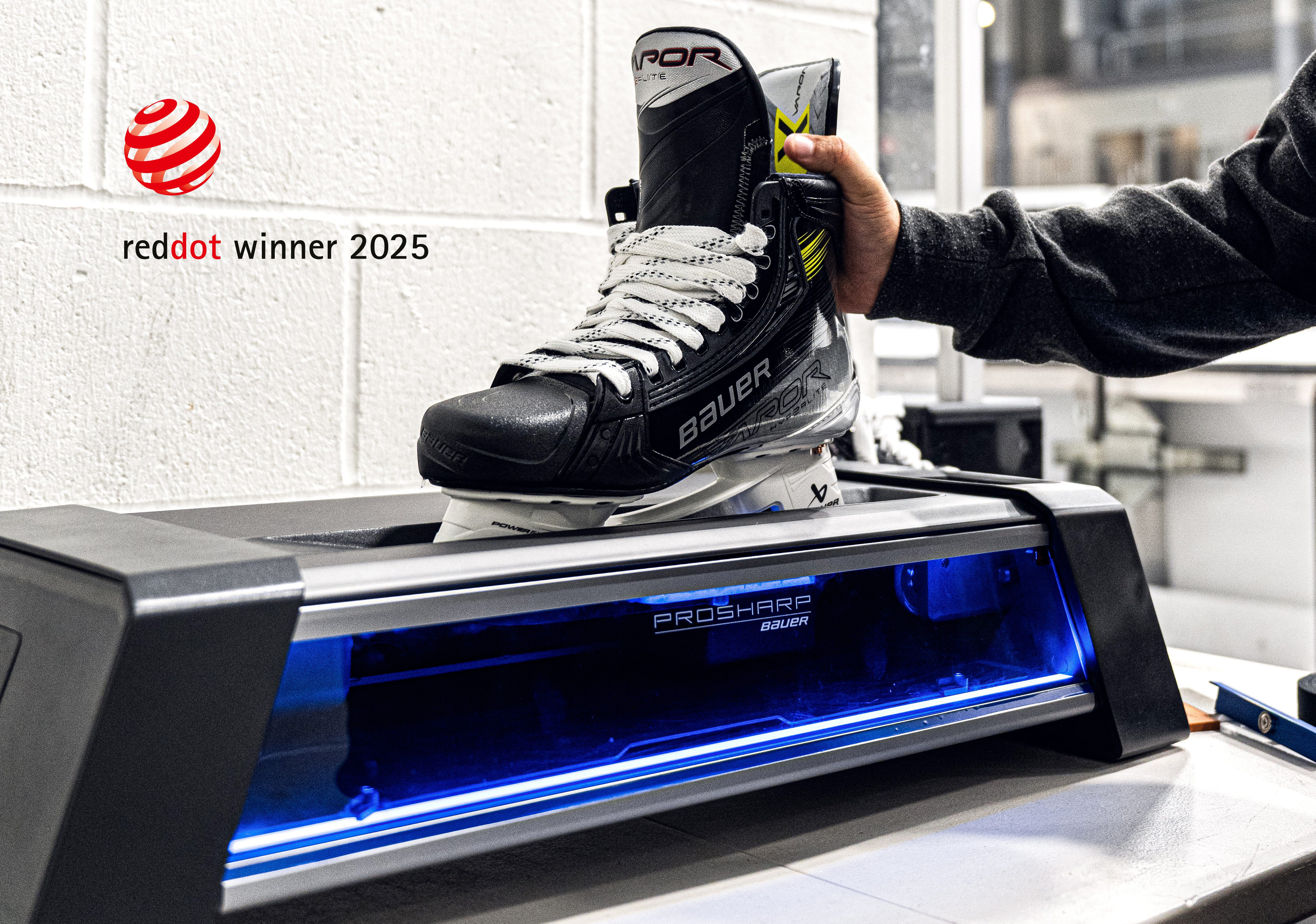 Une personne place un patin de hockey sur glace Bauer noir dans une machine à affûter les patins ProSharp ; le logo du prix Red Dot Winner 2025 est visible sur le mur blanc à l'arrière-plan, reflétant une Stratégie de design au Québec.