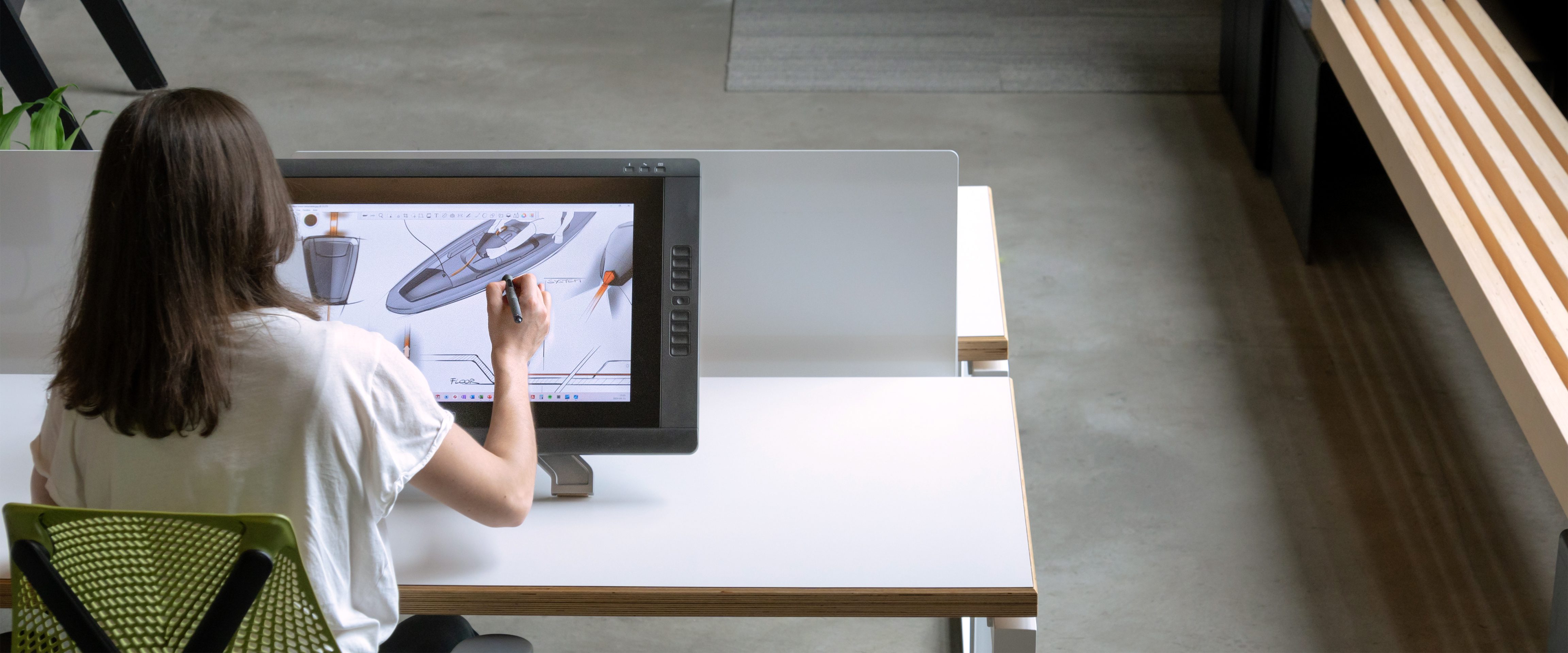 Une personne aux cheveux longs est assise à un bureau dans un espace moderne, utilisant un stylo numérique pour concevoir un modèle de bateau en 3D sur un grand ordinateur à écran tactile dans une agence de développement de produits à Montréal.