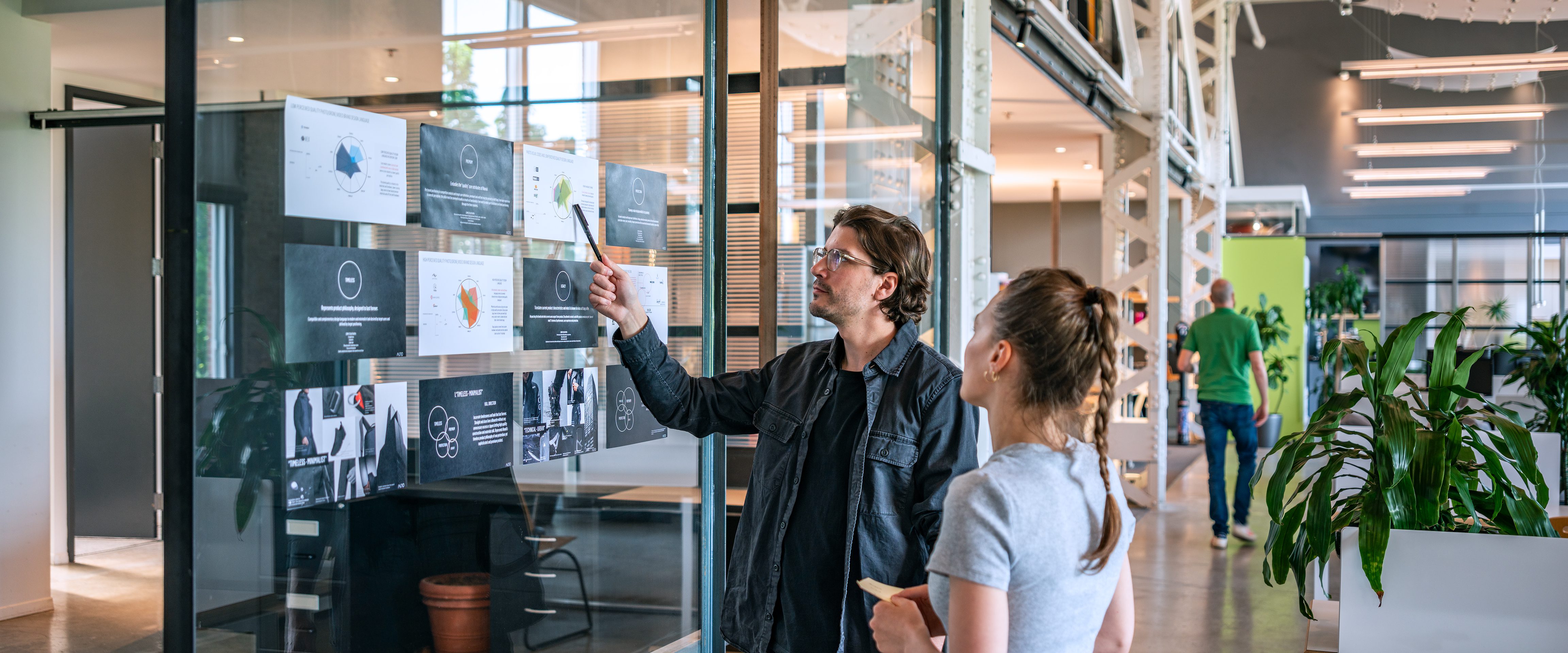 Deux personnes se tiennent dans une Agence de design industriel moderne à Montréal, discutant de tableaux épinglés sur une paroi en verre. L'un pointe du doigt les documents tandis que l'autre écoute, entouré de plantes et d'une atmosphère ouverte et collaborative.