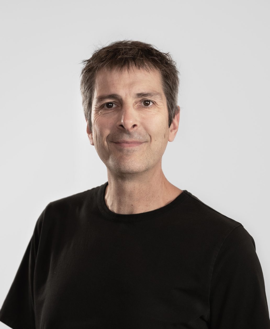 Un homme aux cheveux bruns courts sourit doucement à l'appareil photo. Il porte un simple t-shirt noir et se tient sur un fond gris clair simple, reflétant le style professionnel mais accessible d'une agence de développement de produits à Montréal.