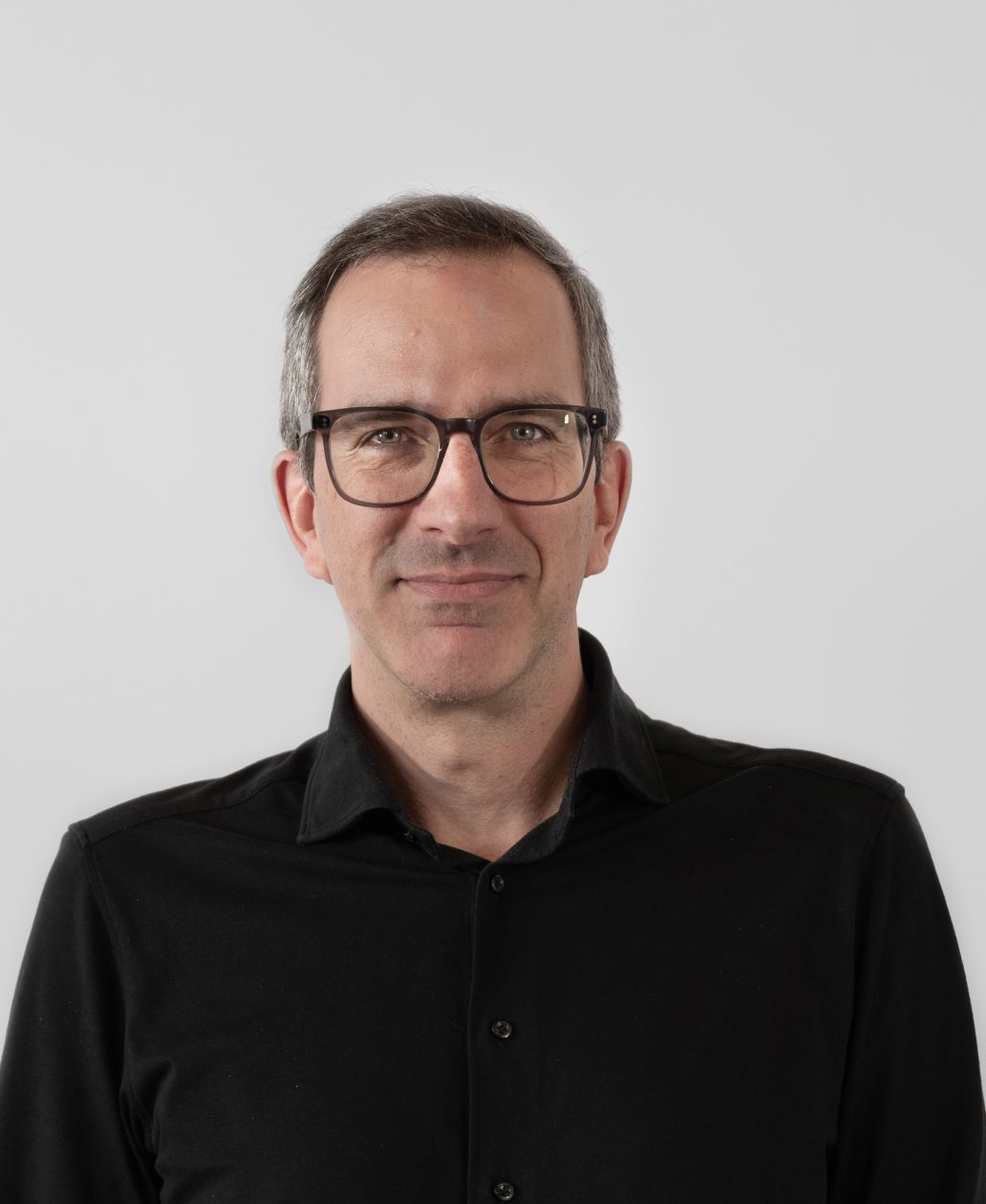Un homme aux cheveux bruns courts, portant des lunettes et un léger sourire, vêtu d'une chemise noire boutonnée, se tient devant un fond blanc uni - une image que l'on voit souvent dans une agence d'ingénierie mécanique à Montréal.