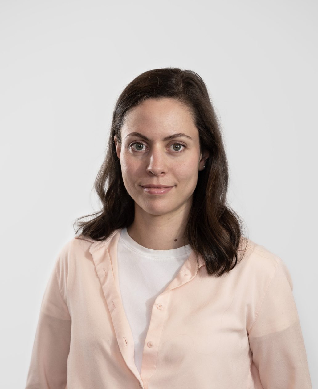 Une femme aux cheveux bruns ondulés, portant une chemise rose pâle boutonnée sur un haut blanc, se tient sur un fond gris clair. Son expression neutre reflète le professionnalisme d'une agence de design industriel à Montréal.