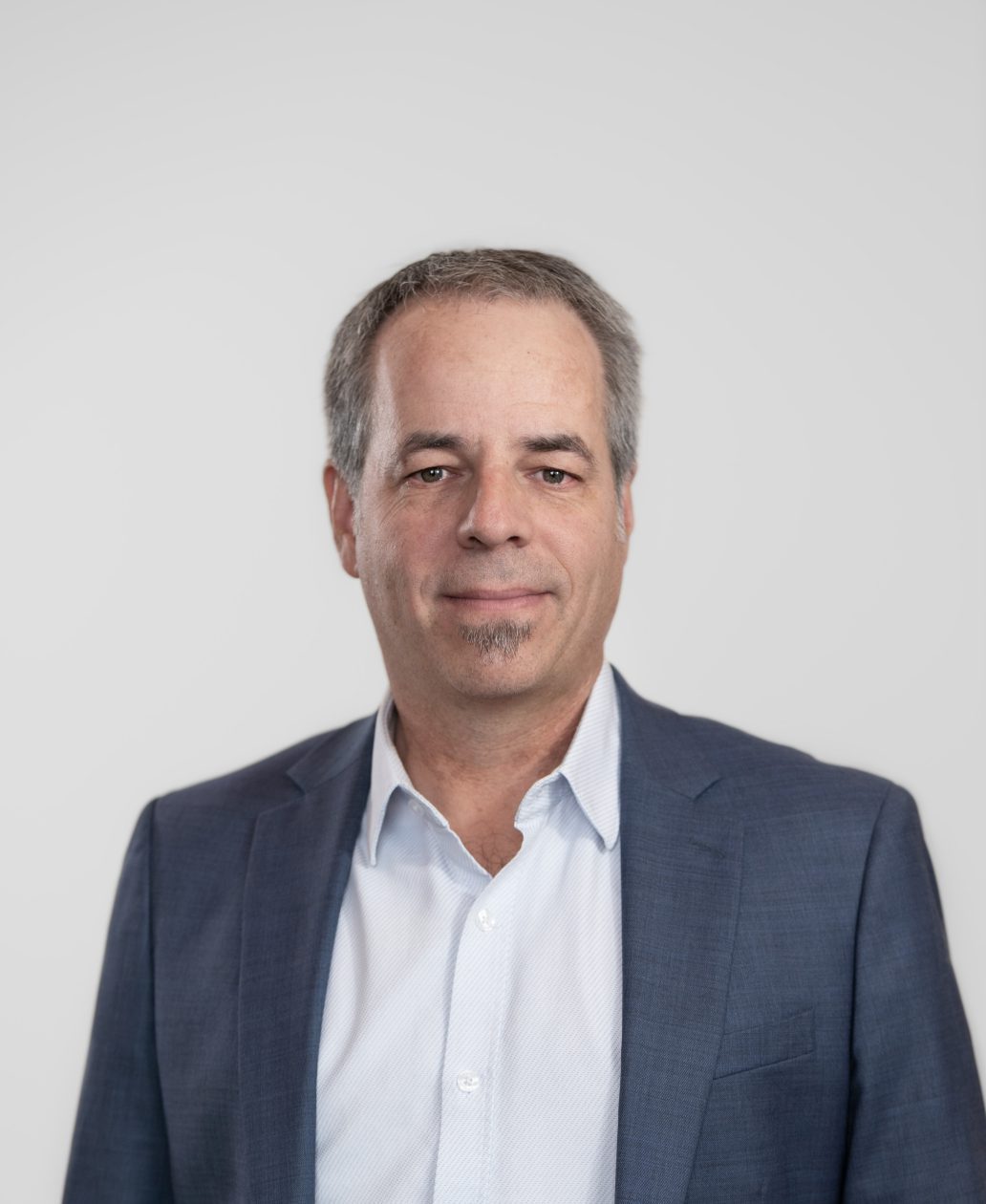 Un homme d'âge moyen aux cheveux gris courts et à la barbichette taillée, vêtu d'une chemise bleu clair et d'un blazer bleu, se tient sur un fond gris clair uni, représentant une agence de design industriel à Montréal avec une expression neutre.