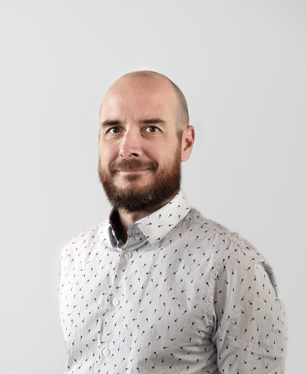 Un homme au crâne chauve et à la barbe fournie, vêtu d'une chemise à boutons à motifs de couleur claire, se tient sur un fond gris clair uni, avec un léger sourire - reflétant l'esprit créatif d'une agence de développement de produits à Montréal.