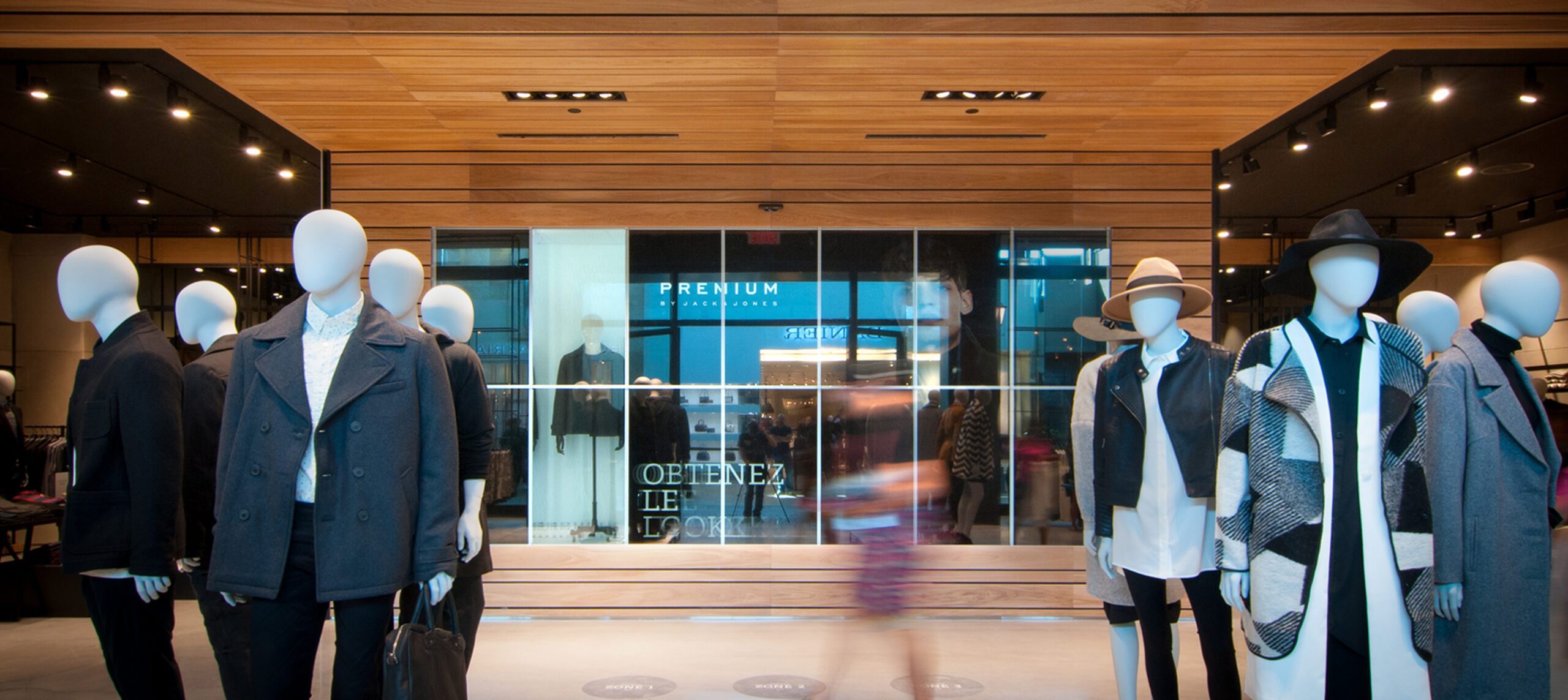 L'entrée d'un magasin de vêtements présente des mannequins portant des tenues modernes et élégantes. Le design épuré s'inspire de la Stratégie de design au Québec, avec des plafonds en bois, des murs en verre et des enseignes visibles lorsque l'on passe devant le magasin.